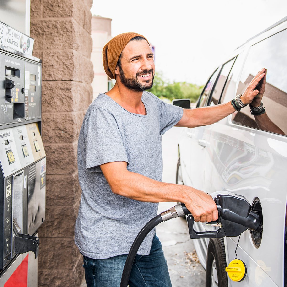 A man pumps gas into a white vehicle at a gas station, smiling while resting his hand on the car, surrounded by a bright outdoor environment.