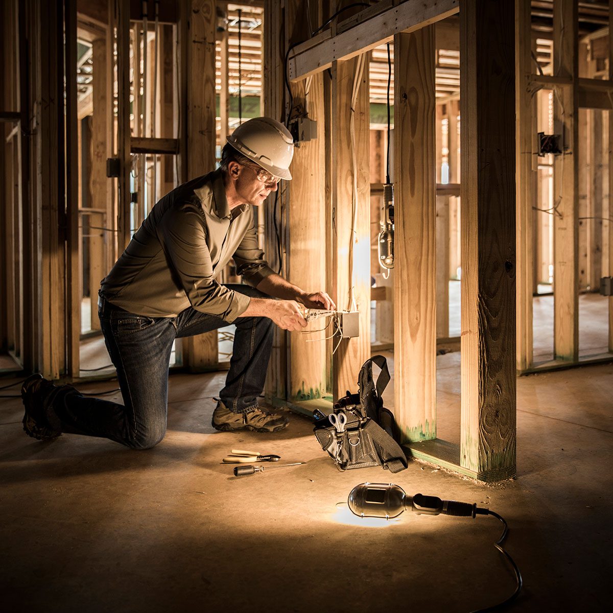 A man kneels, connecting wires inside a partially constructed building, with tools nearby and a lamp illuminating the workspace amidst wooden framing.