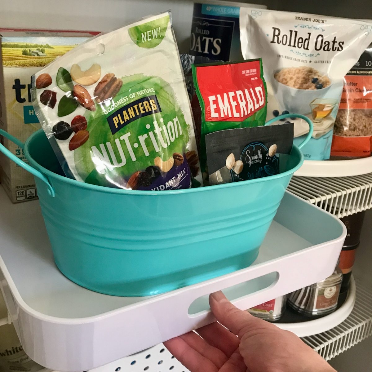 A hand reaches for a white shelf holding a turquoise basket filled with snack packages, surrounded by various food items on organized shelves.