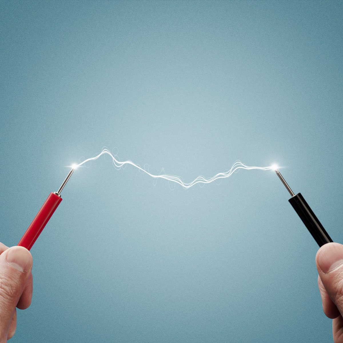 Two hands hold red and black lead wires, connecting with a visible spark or energy discharge, against a soft, light blue background.