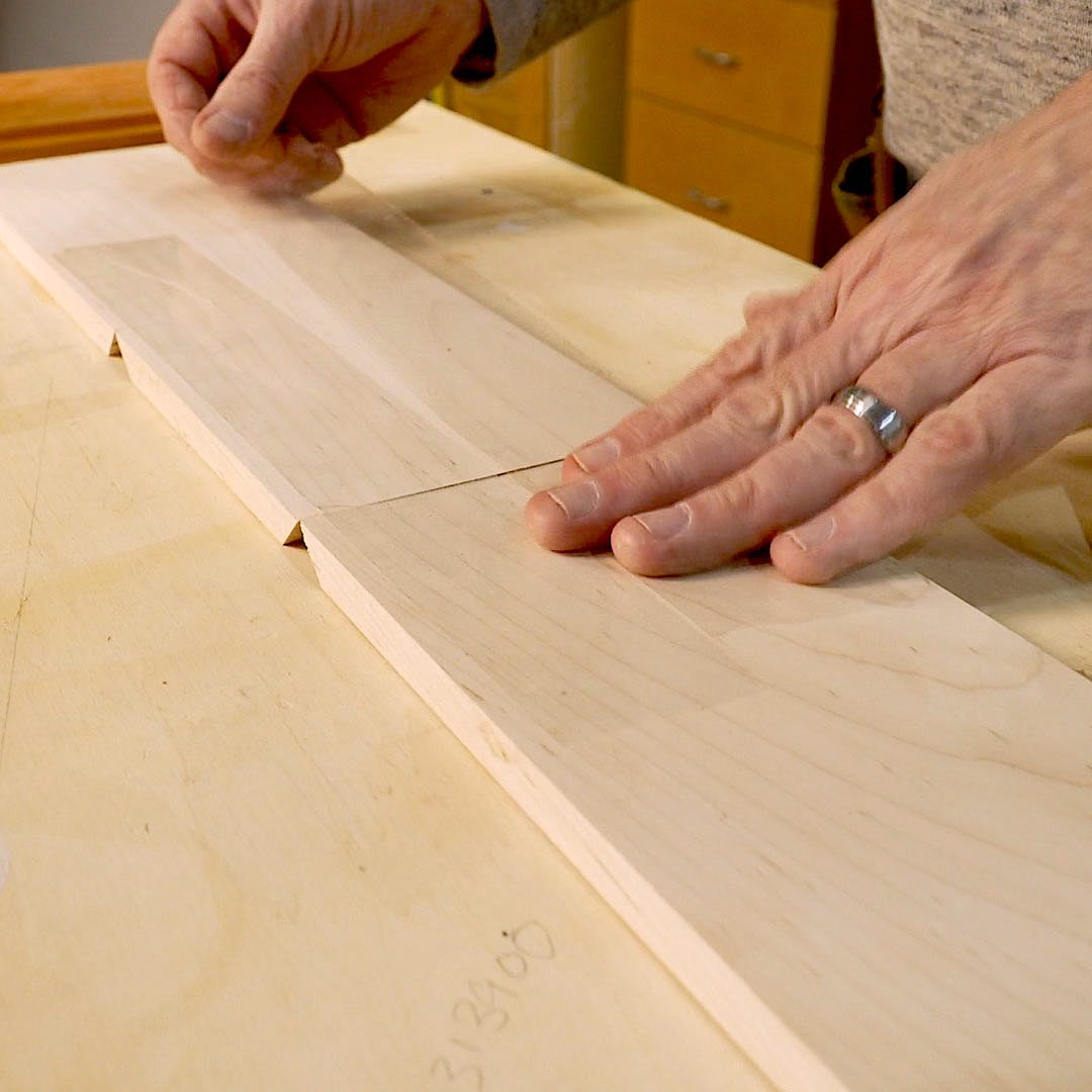 A hand smooths a joint between two wooden pieces on a work surface in a workshop setting, surrounded by tools and cabinets.