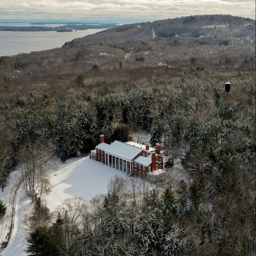 A red brick mansion with white pillars sits surrounded by snow-covered trees, with a lake and hills in the background under a cloudy sky.