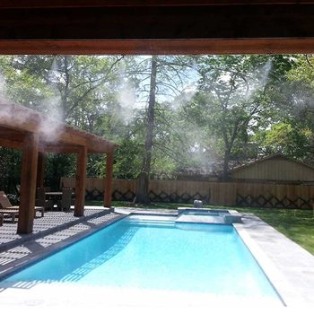 A misting system releases water vapor above a clear swimming pool, surrounded by wooden structures and a lush green landscape under a bright sky.