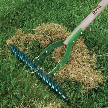 A lawn rake with a spiral metal head is pulling up dry grass clippings, surrounded by lush green grass in a well-maintained yard.