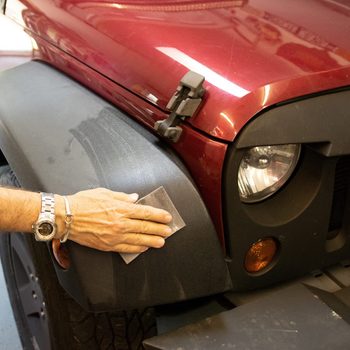 A hand scrubs a black fender of a red vehicle, removing dirt in a well-lit workshop with tools and equipment visible in the background.