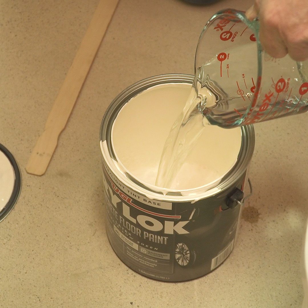 A person pours liquid from a measuring cup into a can of floor paint on a countertop, surrounded by a stirring stick and a clean workspace.