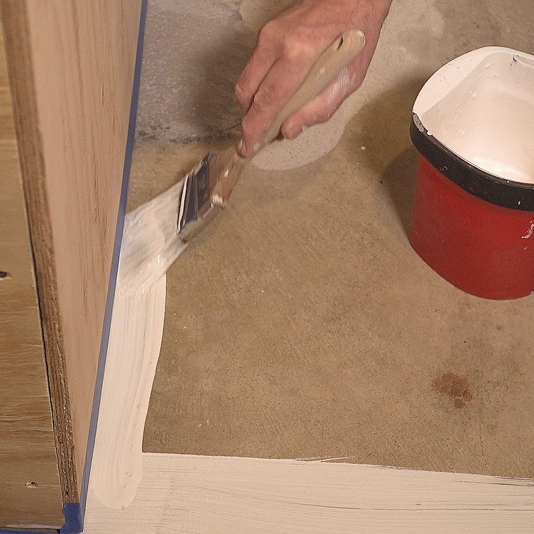 A hand uses a brush to apply white paint to a surface. A red paint bucket rests nearby on a concrete floor.