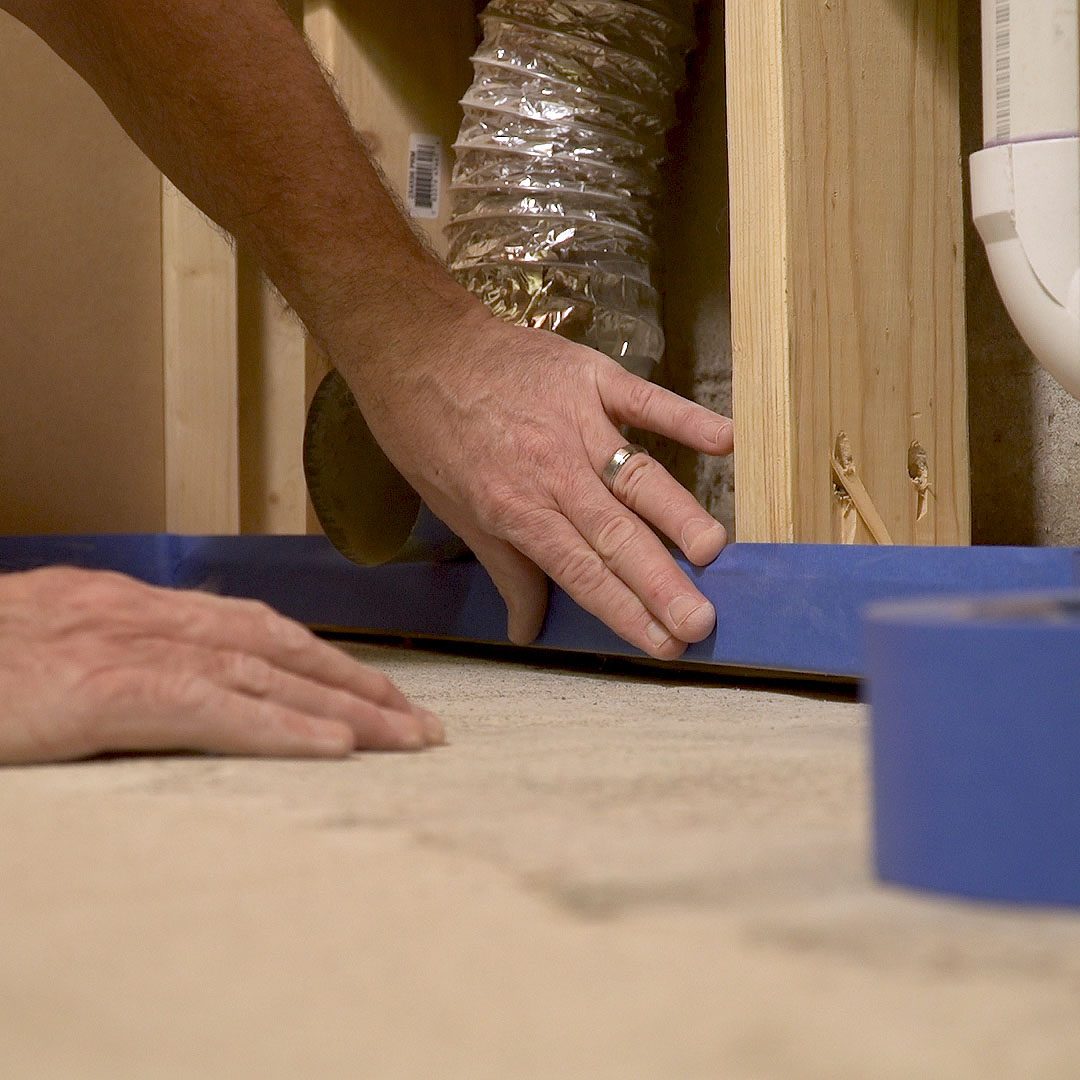A hand is pressing blue tape against the floor, preparing a surface for a project in a room with exposed wooden structures and ducting nearby.
