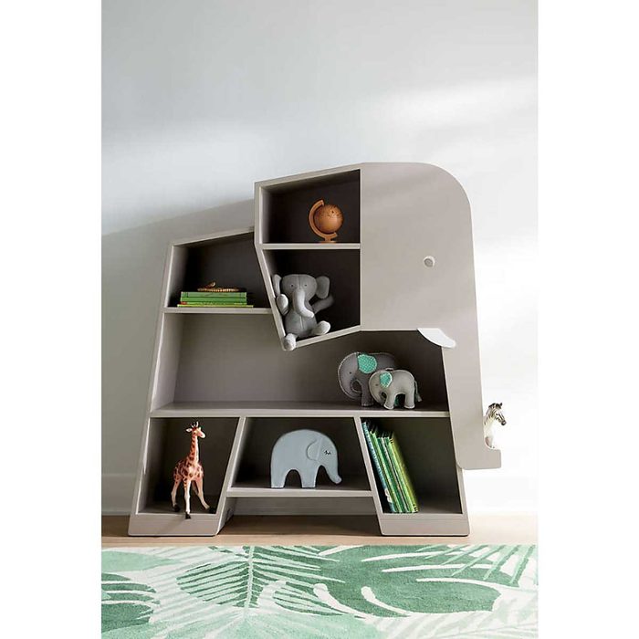 A large, gray elephant-shaped bookshelf displays various animal figurines and books in a bright, minimalist room with a patterned green rug.