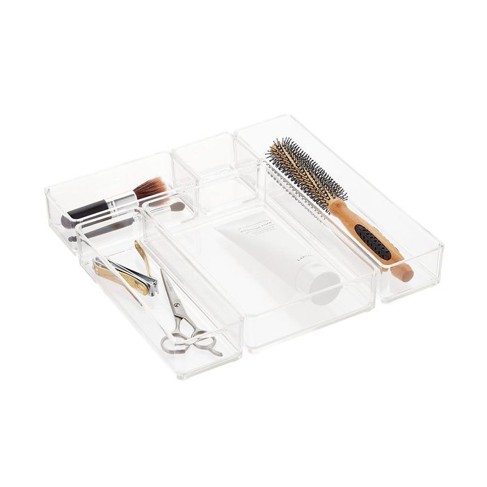 A clear tray holds various beauty tools: brushes, scissors, a hairbrush, and a cream tube, organized in separate compartments, on a plain surface.