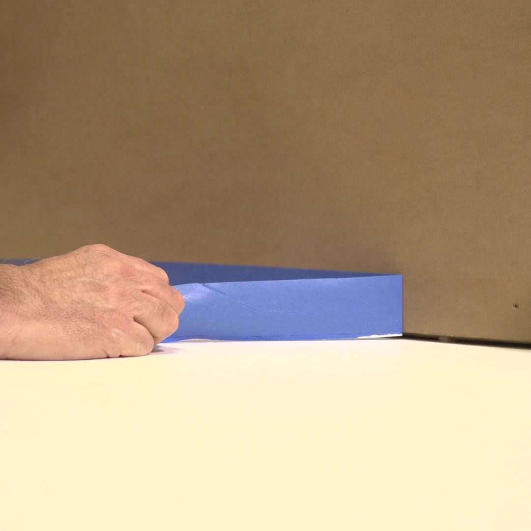 A hand adjusts a piece of blue tape along the edge of a surface, with a plain brown background behind it.