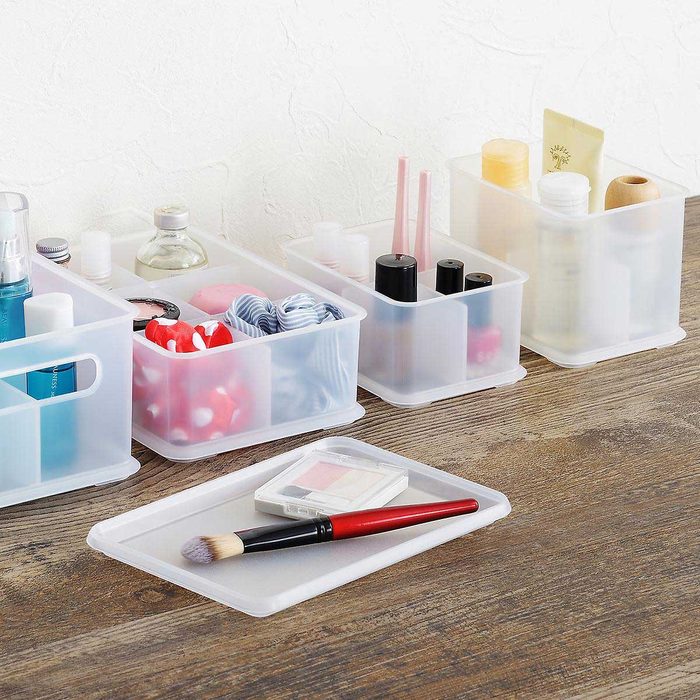 Four transparent storage containers hold various cosmetics and bathroom items on a wooden surface, with a makeup brush and palette resting beside them.