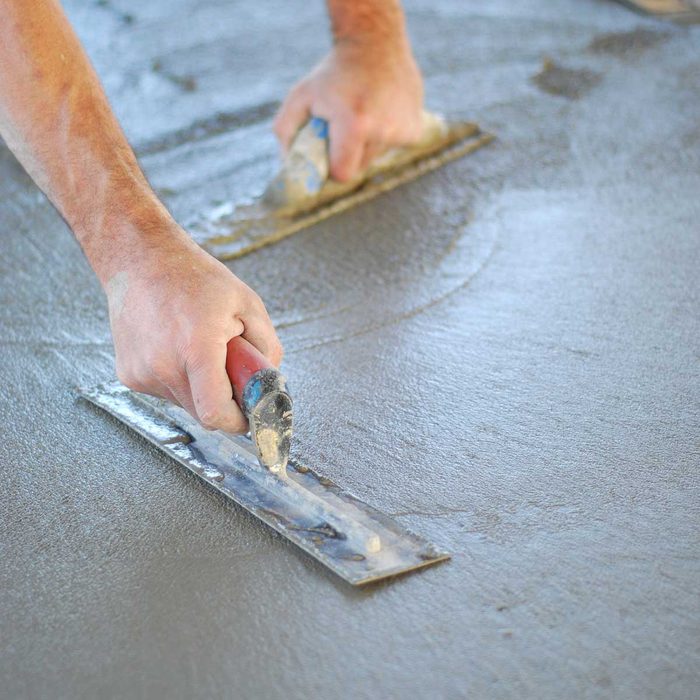 Hands are smoothing fresh concrete with trowels, creating a flat surface on a partially cured, wet floor.