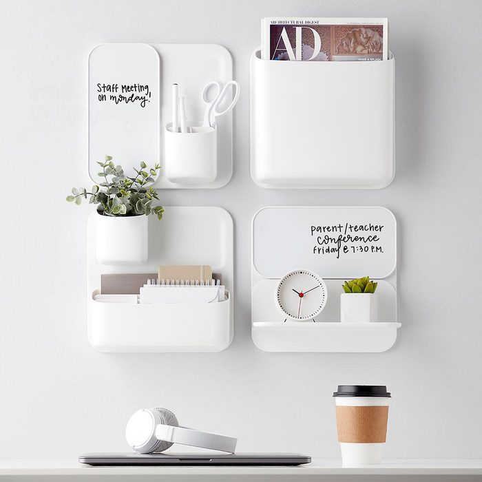 Four white wall-mounted organizers hold various items like plants, scissors, paperwork, and a clock, set against a light wall with a coffee cup below.
