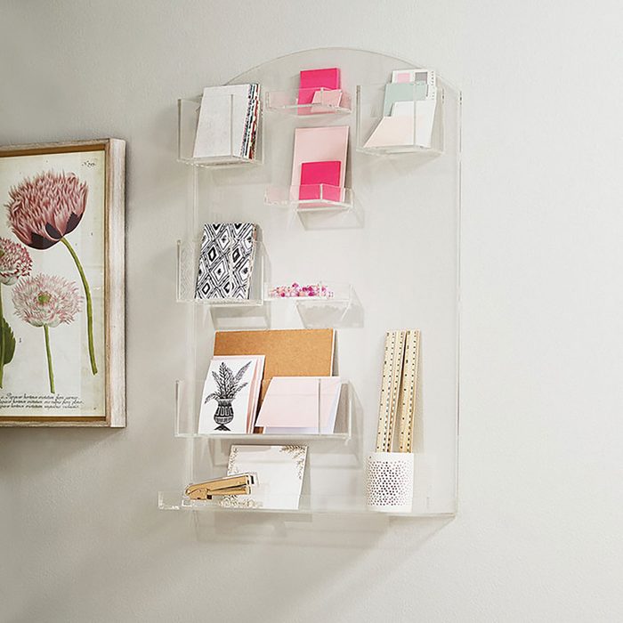 A wall-mounted display holds various cards and stationery items, organized in clear holders, with a floral artwork to the left against a light wall.