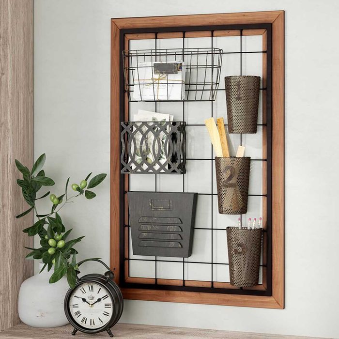 A wall-mounted organizer displays various compartments holding papers and writing tools, beside a clock and greenery on a wooden surface.
