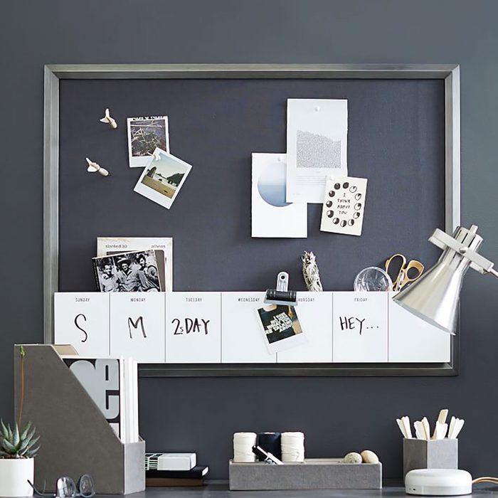 A framed bulletin board displays pinned photos and notes, while a calendar lists days. A stylish lamp and storage containers sit below on a desk.