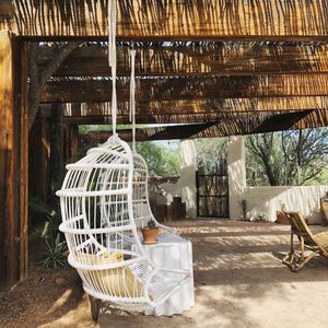 A white swing chair hangs from a wooden structure, inviting relaxation under shaded patterns, surrounded by a rustic patio and greenery.