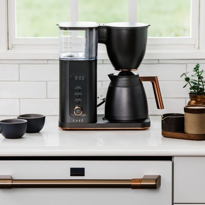 A coffee maker brews coffee, positioned on a countertop with three dark cups. Sunlight illuminates a modern kitchen with green views outside.