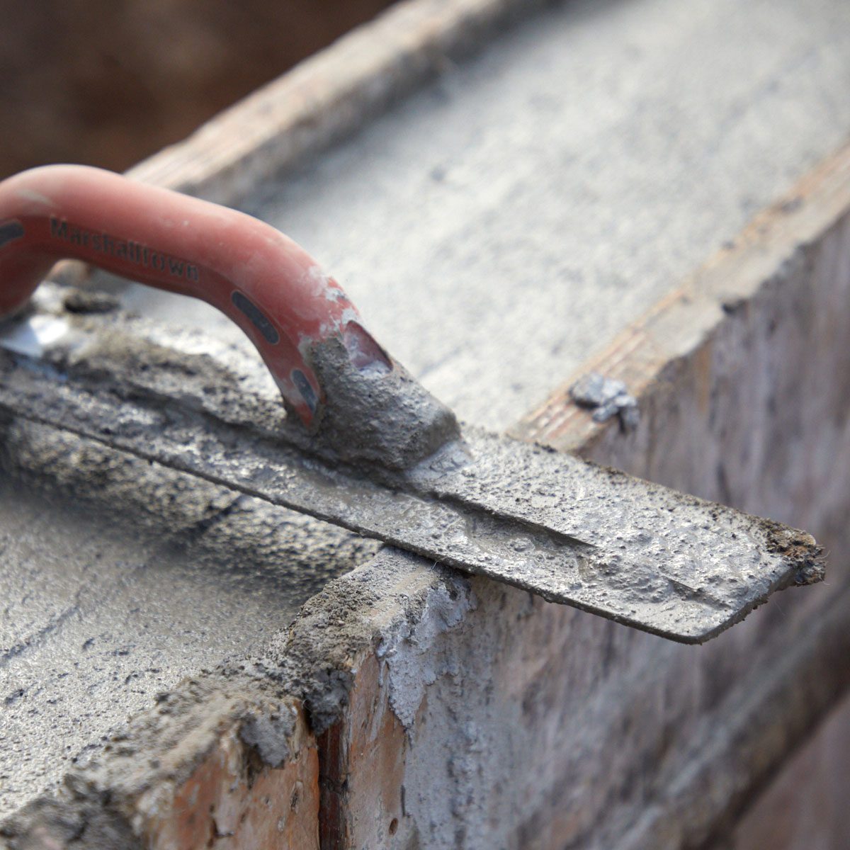 A trowel is smoothing wet concrete on a block surface, surrounded by unfinished construction materials, indicating ongoing masonry work.