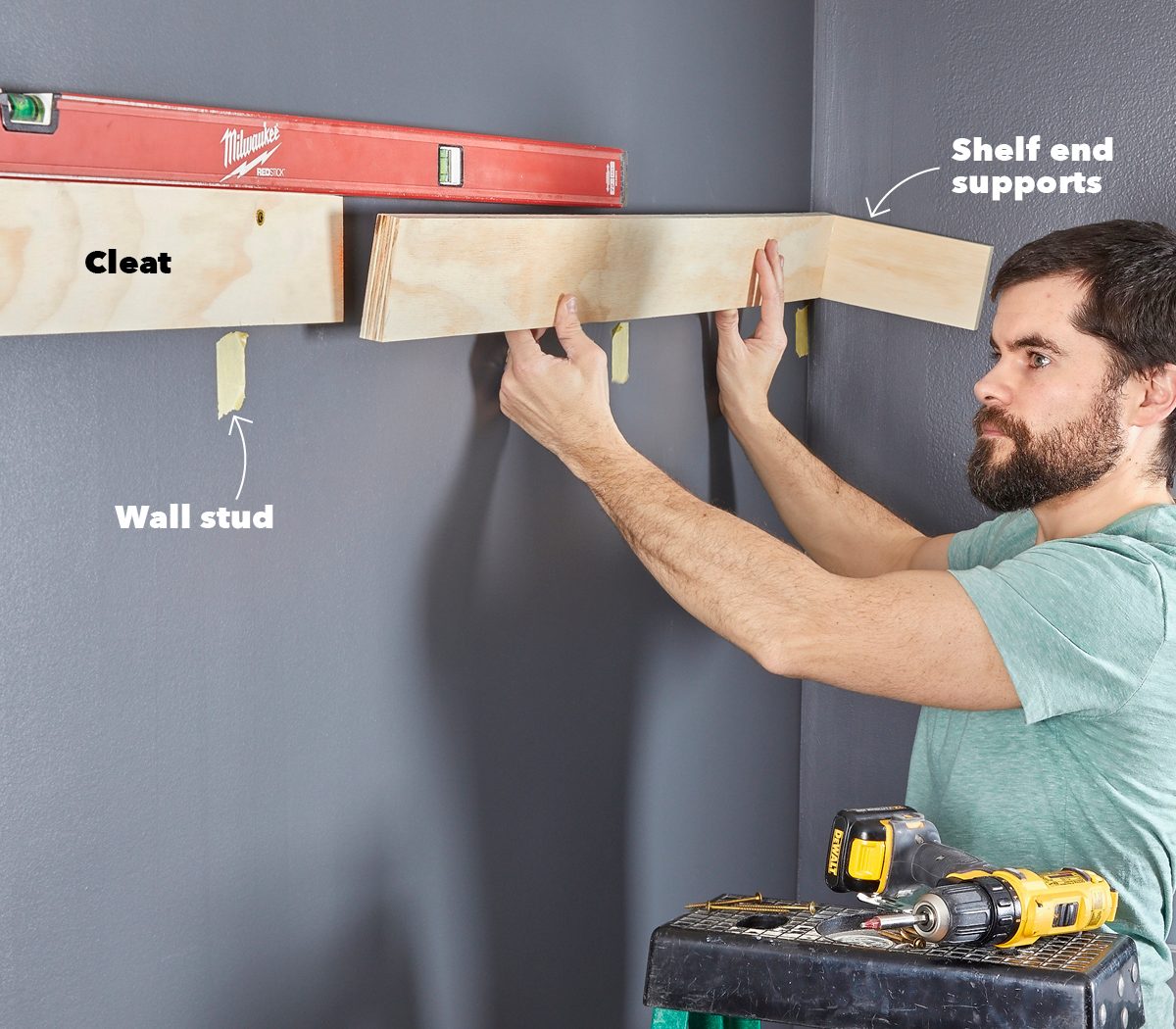 A man installs a wooden shelf using a level and supports, positioning it against a dark wall, with tools on a nearby step stool.