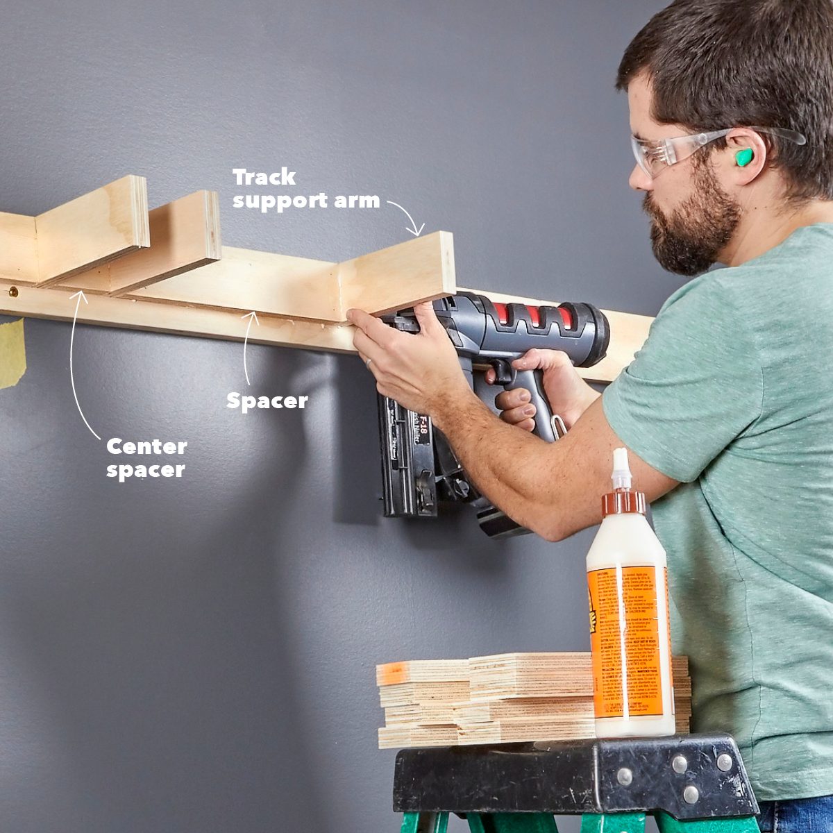 A man uses a nail gun to attach wooden support components to a wall, while standing on a ladder beside a stack of wood and glue.