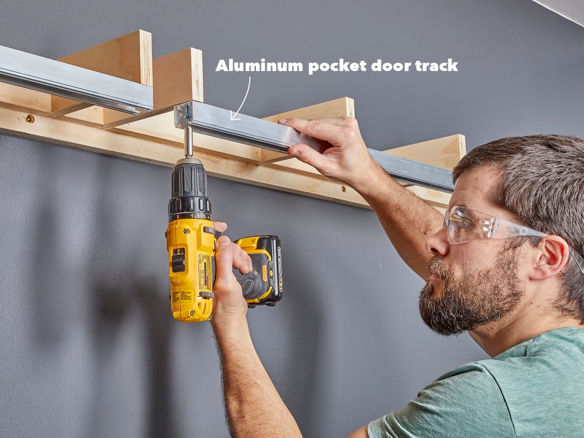 A person drills into an aluminum pocket door track mounted on a wooden structure against a dark gray wall while wearing safety goggles.