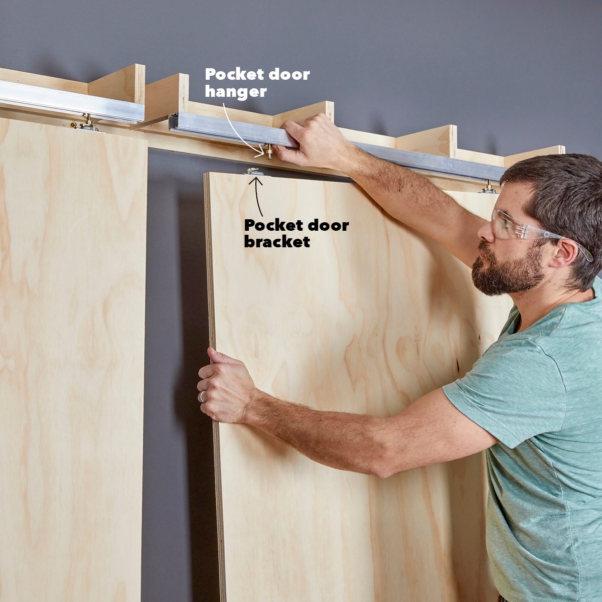 A person installs a pocket door bracket onto wooden panels, adjusting it beneath a horizontal track mounted on a gray wall.