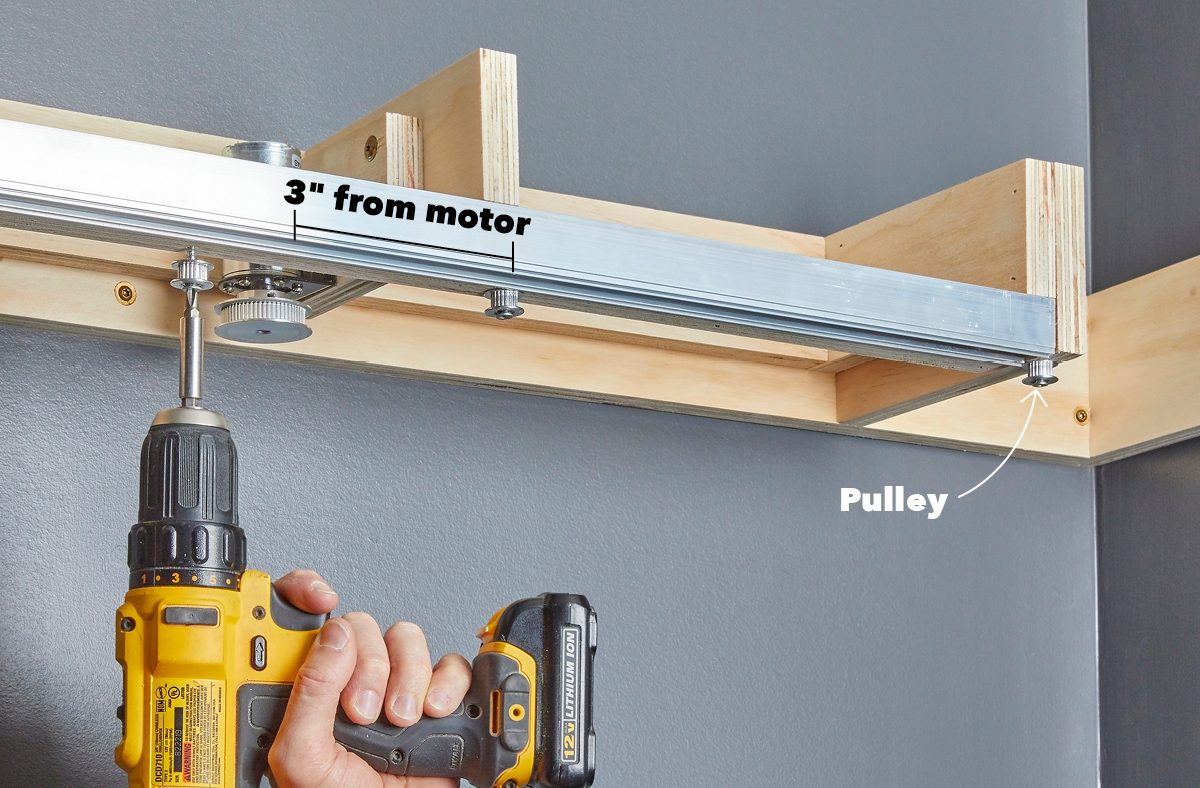 A hand holds a drill attaching a metal rail with a pulley to a wooden frame mounted on a gray wall.