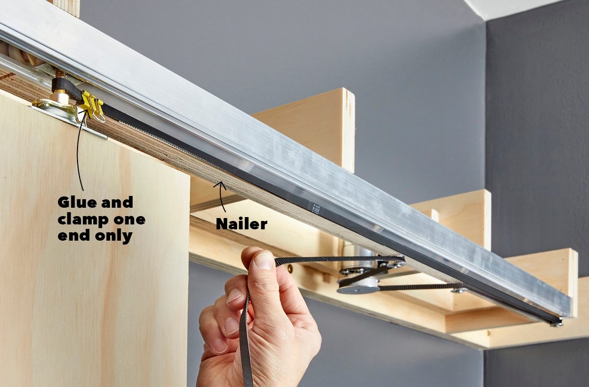 A hand holds a strap, securing it to a metal guide, while wooden structures are visible in a gray room.