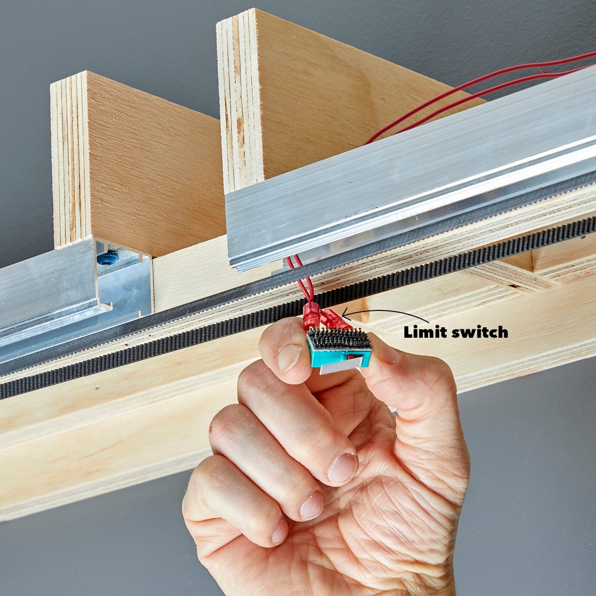 A hand holds a limit switch connected by wires, positioned near a wooden structure with metal components, suggesting installation or maintenance within a mechanical system.
