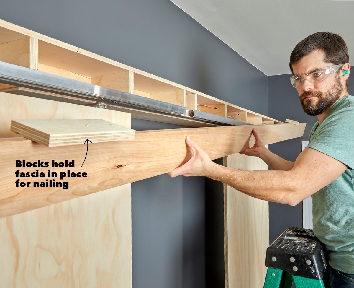 A man holds a wooden fascia in place, preparing to nail it against walls that have a dark gray paint, using blocks for support.