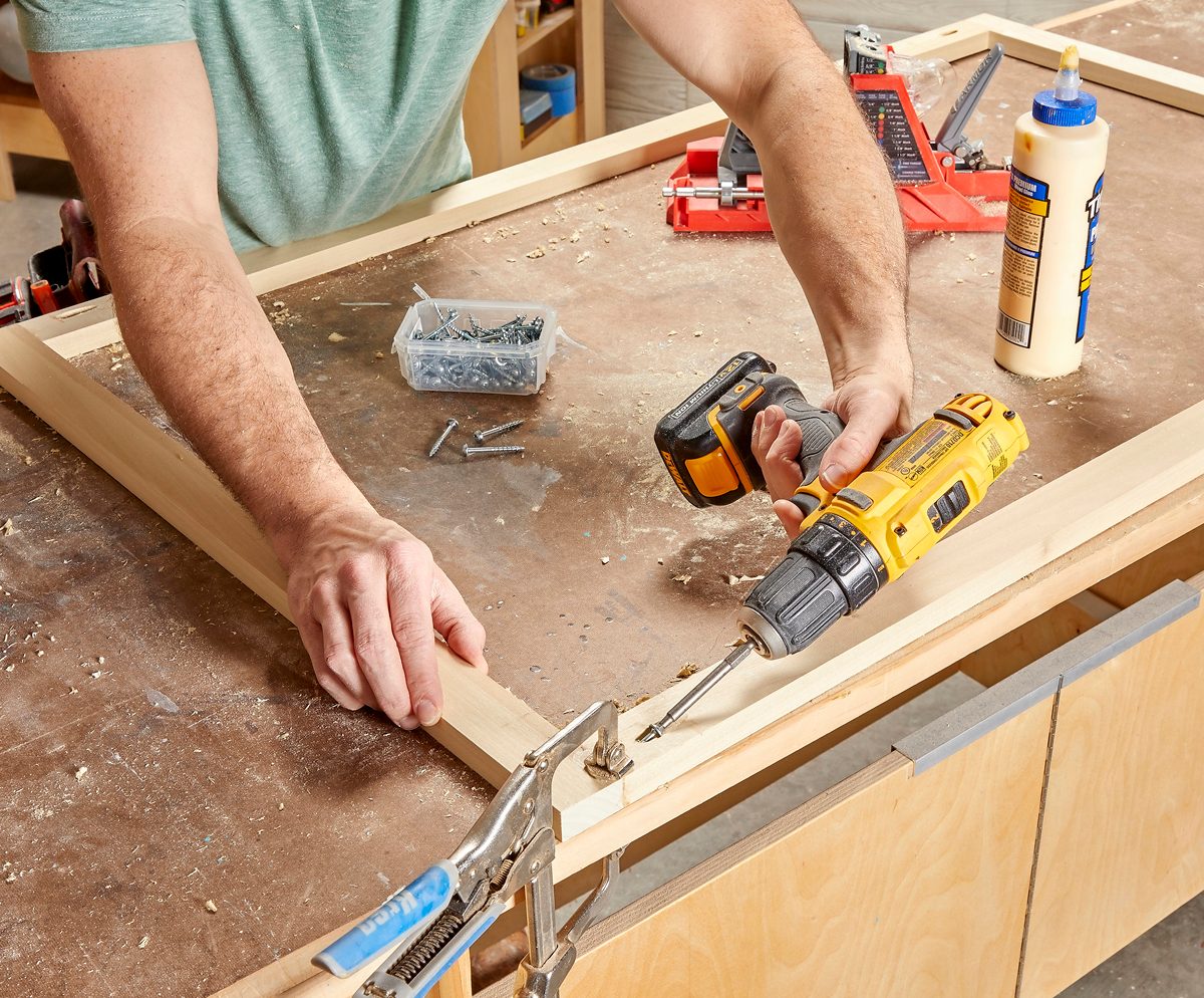 A person uses a power drill to secure wooden planks together on a workbench, surrounded by tools and a container of screws.