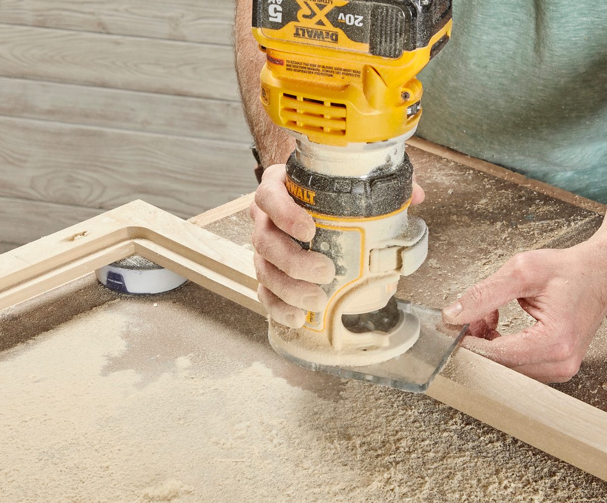 A person uses a handheld router on a wooden piece, creating a smooth edge, surrounded by wood shavings on a work surface.