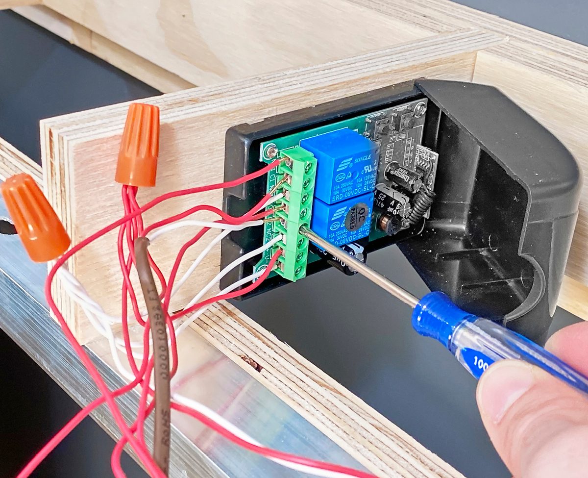 A person uses a screwdriver to connect wires to a circuit board mounted on a wooden frame, surrounded by several loose electrical wires.