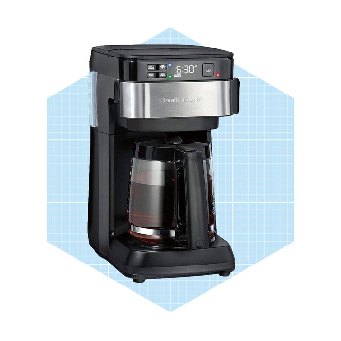 A black and silver coffee maker brews coffee, positioned on a blue grid background, showcasing its digital display and glass carafe.