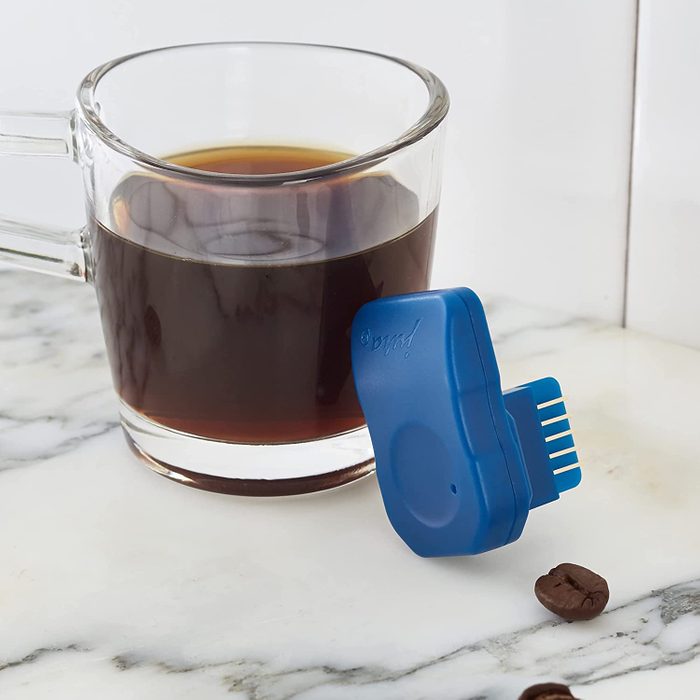 A blue coffee tool rests beside a clear cup of dark coffee on a marble surface, with a few scattered coffee beans nearby.