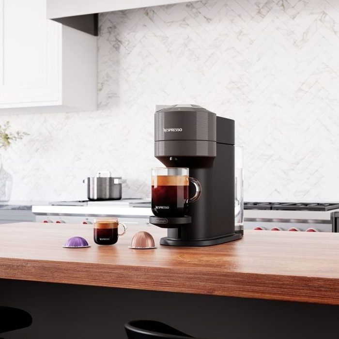 A coffee machine stands on a wooden countertop, brewing coffee into a glass cup, with two coffee capsules beside it in a modern kitchen setting.
