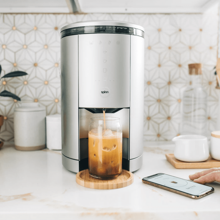 A sleek coffee machine dispenses iced coffee into a glass on a wooden coaster, while a smartphone nearby displays a control interface on a kitchen counter.