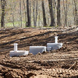 Three concrete septic tank covers are positioned in a cleared, uneven dirt area, surrounded by sparse trees and a dirt pathway in the background.