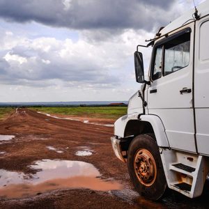 A white truck is parked on a muddy dirt road, reflecting puddles beneath it, with a cloudy sky and green fields in the background.