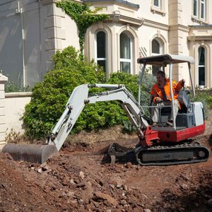 A small excavator digs into soil while a man in an orange jacket operates it near a lush garden and a large, ornate building.