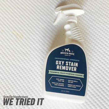 A spray bottle of Oxy Stain Remover is positioned on a textured surface, indicating a cleaning product designed for various stains.