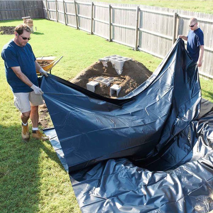 Two men are unfolding a large black tarp over a dirt mound in a backyard, surrounded by grass and wooden fencing.