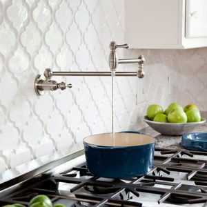 A polished faucet streams water into a blue pot on a stove, set against a white tiled backsplash and surrounded by green apples on a countertop.