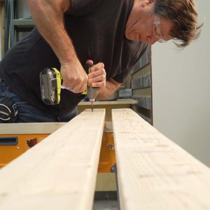 A man uses a power drill to fasten two wooden planks together, focused intently in a workshop environment with shelves in the background.