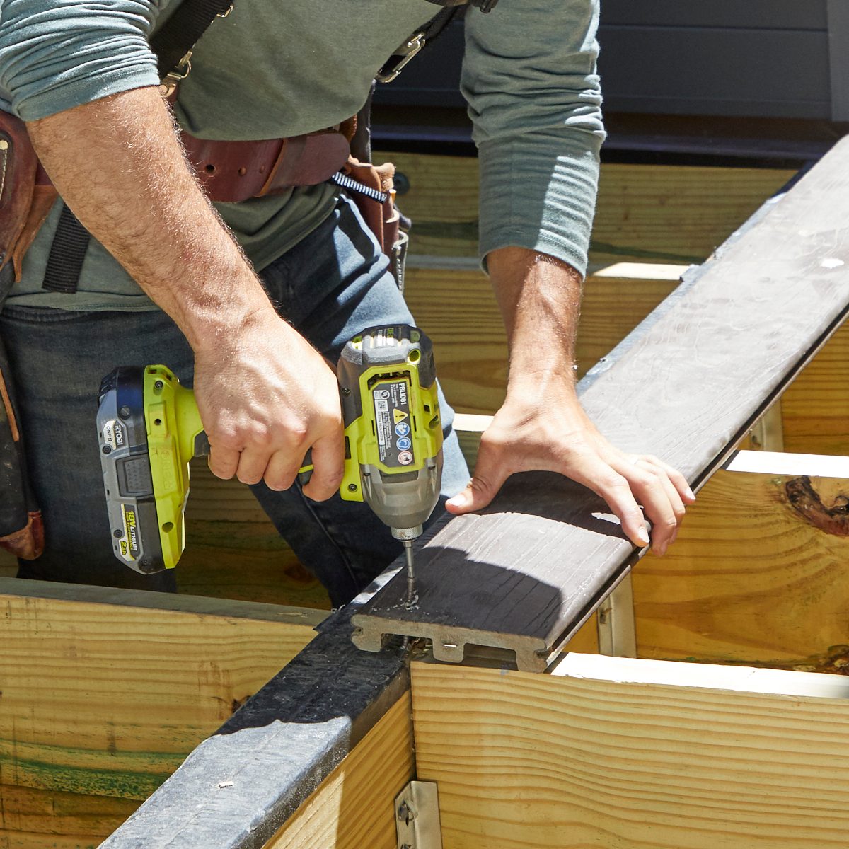 A person uses a power drill to secure a dark board onto a structure made of wooden beams, working in a brightly lit outdoor environment.