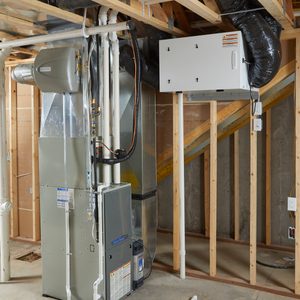 A heating unit stands in an unfinished basement, connected to ducts and pipes, with wooden framing and a staircase visible in the background.