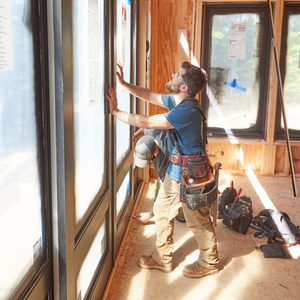 Two construction workers are installing windows in a wooden-framed room, focused on precise measurements amidst tools and materials scattered on the floor.
