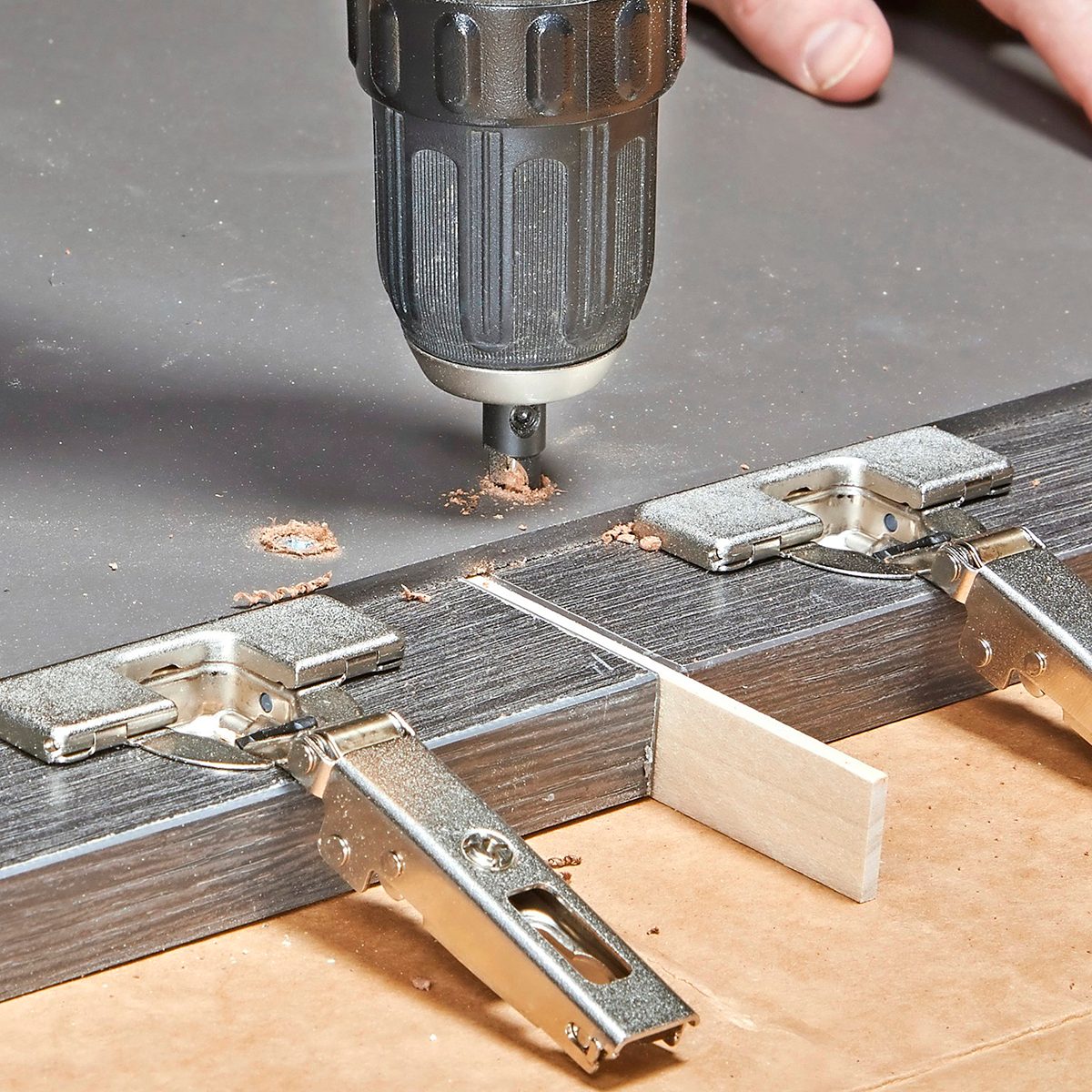 A drill is making a hole in a dark board secured by metal clamps, with wood shavings visible on the surface, suggesting woodworking activities.